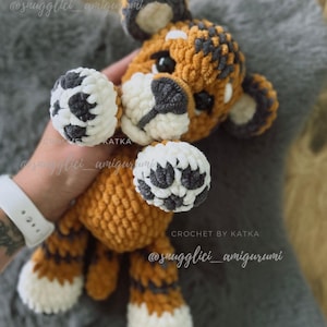 May include: A handmade crochet tiger amigurumi in orange, white, and gray. The tiger has a textured body, white paws, and a gray and white face. The text "CROCHET BY KATKA" and "@snugglici_amigurumi" are visible.