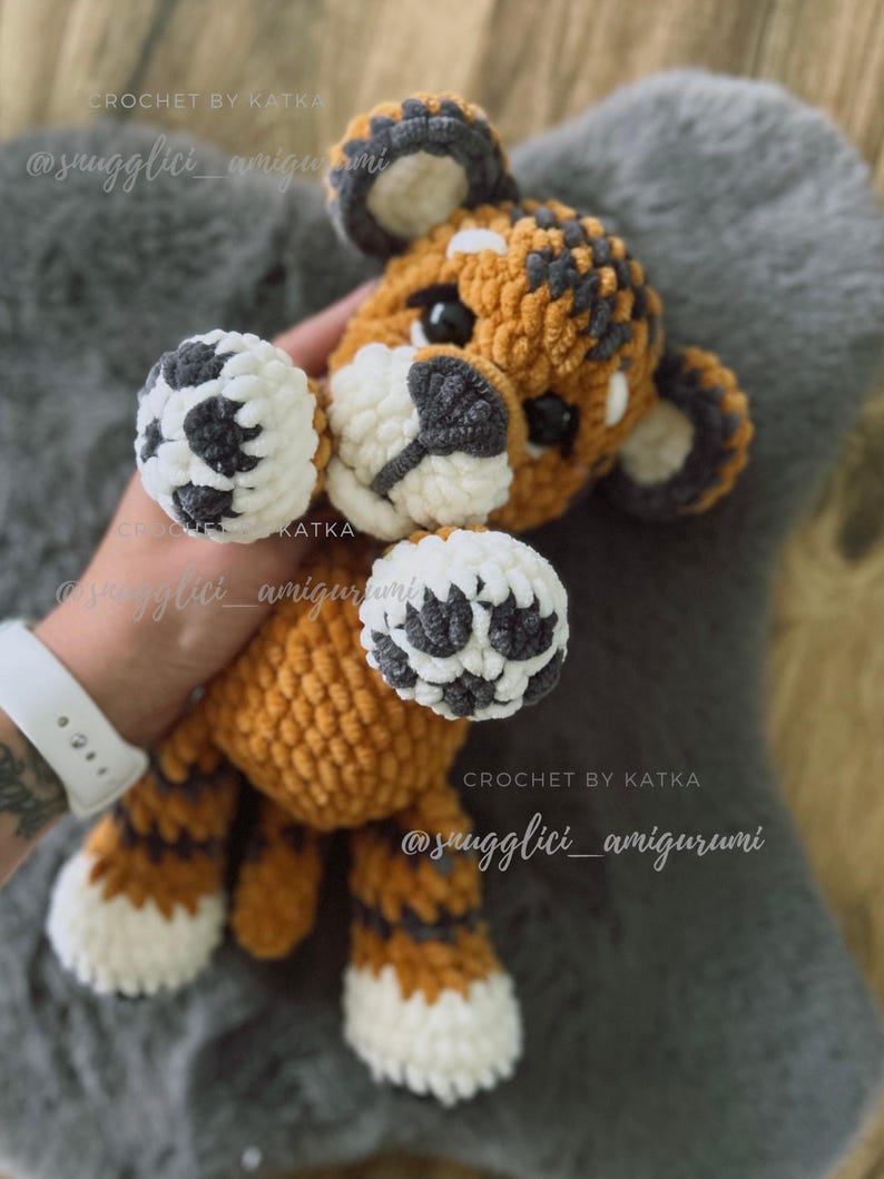 May include: A handmade crochet tiger amigurumi in orange, white, and gray. The tiger has a textured body, white paws, and a gray and white face. The text "CROCHET BY KATKA" and "@snugglici_amigurumi" are visible.