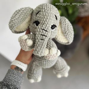 May include: A handmade gray crochet elephant amigurumi with white accents on the ears and feet. The elephant has black eyes and a trunk. The text "CROCHET BY KATKA @snugglici_amigurumi" is visible.