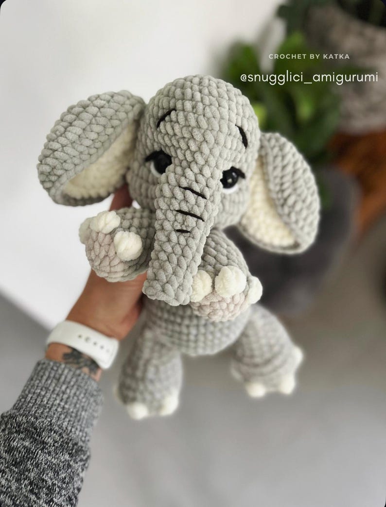 May include: A handmade gray crochet elephant amigurumi with white accents on the ears and feet. The elephant has black eyes and a trunk. The text "CROCHET BY KATKA @snugglici_amigurumi" is visible.
