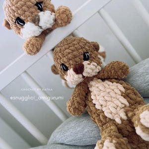 Pu&ograve; includere: Due peluche di lontra all'uncinetto fatti a mano, nei toni del marrone e del crema. Le lontre hanno occhi neri e naso marrone chiaro. Una lontra &egrave; posizionata come se sbirciasse sopra una sponda bianca, mentre l'altra riposa su una superficie grigia. Il testo "CROCHET BY KATKA" e "@snugglci_amigurumi" sono visibili.