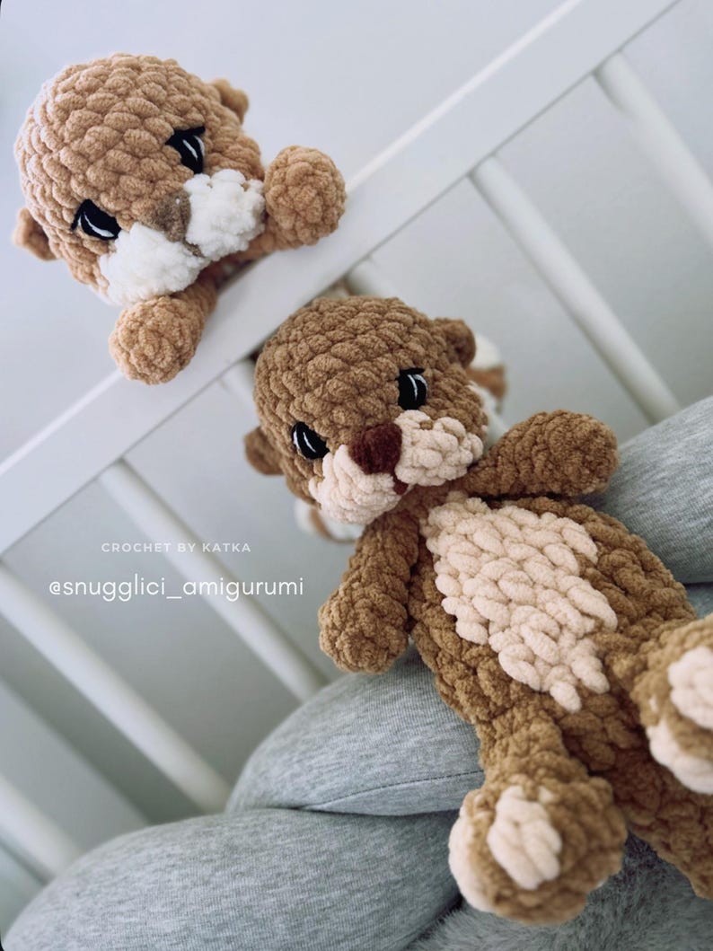 Pu&ograve; includere: Due peluche di lontra all'uncinetto fatti a mano, nei toni del marrone e del crema. Le lontre hanno occhi neri e naso marrone chiaro. Una lontra &egrave; posizionata come se sbirciasse sopra una sponda bianca, mentre l'altra riposa su una superficie grigia. Il testo "CROCHET BY KATKA" e "@snugglci_amigurumi" sono visibili.