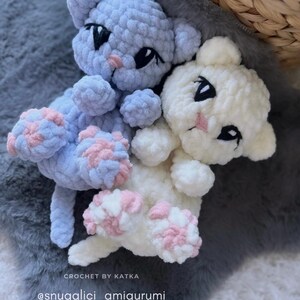 May include: Two handmade crochet cat amigurumi toys. One is light blue, the other is white. Both have black eyes, pink noses, and pink and blue paw pads. The text "CROCHET BY KATKA" and "@snugglici_amigurumi" are visible.