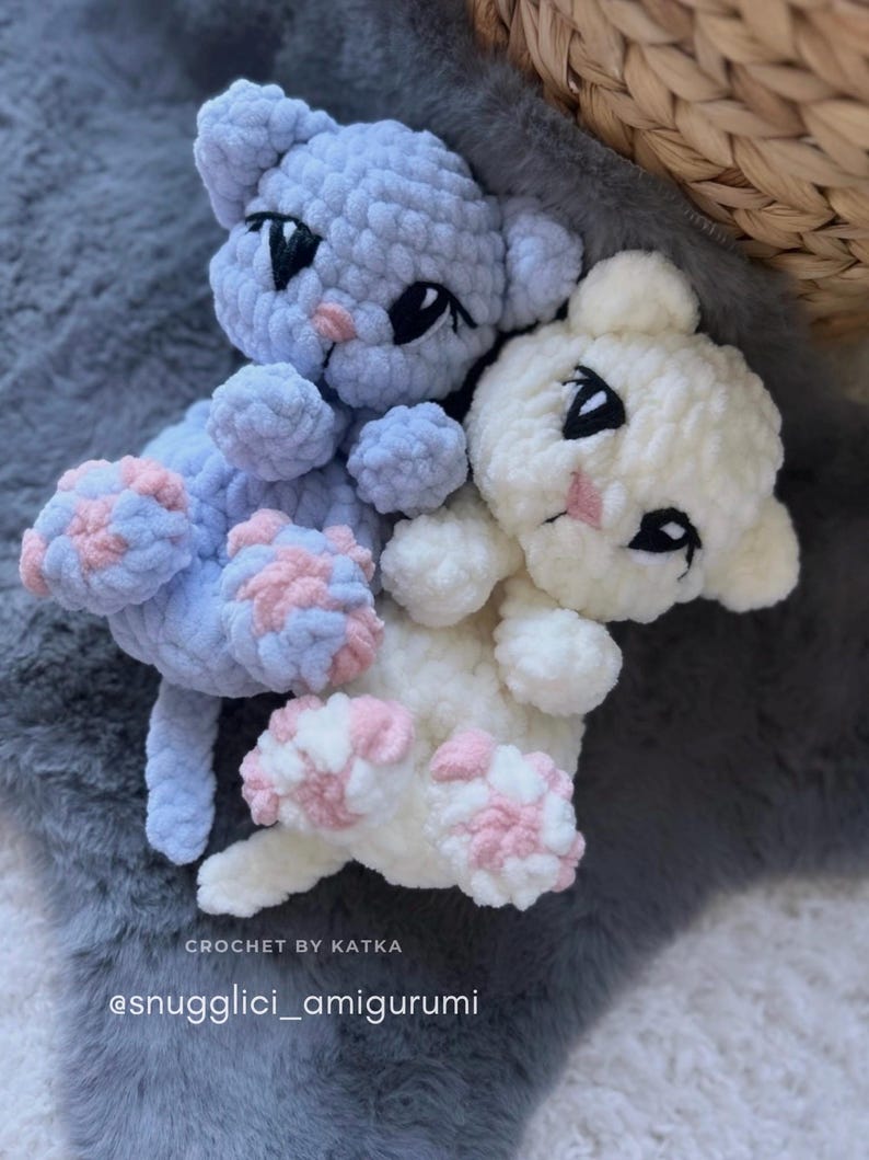 May include: Two handmade crochet cat amigurumi toys. One is light blue, the other is white. Both have black eyes, pink noses, and pink and blue paw pads. The text "CROCHET BY KATKA" and "@snugglici_amigurumi" are visible.