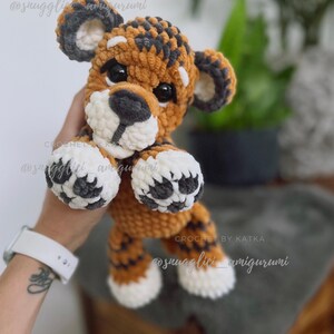 May include: A handmade crochet tiger amigurumi in shades of orange, brown, gray, and white. The plush toy has a round head, small ears, and black eyes. The text "CROCHET BY KATKA" and "@snugglivi_amigurumi" are visible.