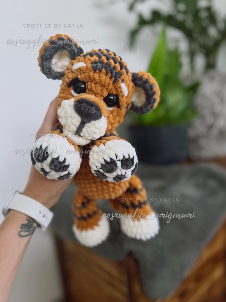 May include: A handmade crochet tiger amigurumi in shades of orange, brown, gray, and white. The plush toy has a round head, small ears, and black eyes. The text "CROCHET BY KATKA" and "@snugglivi_amigurumi" are visible.