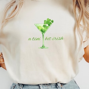 May include: Cream-colored t-shirt with a green martini glass graphic and the text "a tini bit irish." The martini glass is filled with green liquid and topped with shamrocks. The shirt is made of a soft material.