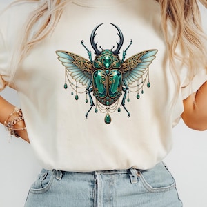 May include: Cream-colored t-shirt featuring a detailed illustration of a jeweled beetle. The beetle has a teal body, ornate gold wings, and dangling green gems. The design is centered on the shirt, creating a striking visual.