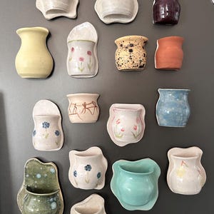May include: A collection of handmade ceramic wall planters in various shapes, sizes, and colors. The planters include a yellow vase, a speckled pot, and several with floral designs. The color palette includes white, blue, orange, and green.
