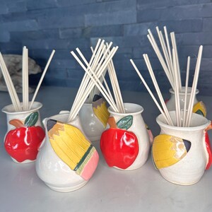 May include: A collection of five ceramic reed diffusers, each featuring a cream-colored base decorated with a raised, hand-painted red apple or yellow pencil design. Each vessel contains a bundle of thin, light-colored wooden diffuser reeds.