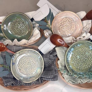 Puede incluir: Cuatro ralladores de cerámica en varios colores, cada uno en una cesta de mimbre con una cuchara de madera, una espátula blanca y un paño. Los ralladores tienen una superficie texturizada. Las hojas decorativas añaden un toque natural.