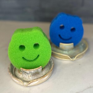 May include: Two smiley face sponges, one green and one blue, sit on ceramic soap dishes. The sponges have a simple face design with two eye holes and a curved smile. The dishes are light-colored with a subtle, organic shape.