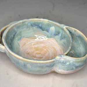 Op de afbeelding: Een handgemaakte keramische kom met een unieke, organische vorm. De kom heeft een lichtblauwe en beige glazuur, waardoor een gemarmerd effect ontstaat. Het heeft een kleine, aangehechte zijkom en twee handvatten. Ideaal om voedsel in te serveren of als decoratief stuk.