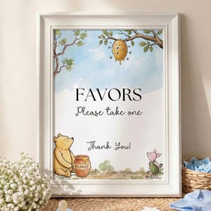 May include: A framed print with a Winnie the Pooh theme. The print features a watercolor illustration of Winnie the Pooh, Piglet, and a honey pot. The text on the print reads "FAVORS Please take one Thank You!" A baby blue onesie and socks are next to the print.