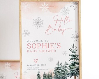 以下が含まれることがあります： 冬をテーマにした、額入りのベビーシャワーのサイン。「Hello Baby」と「Sophie's Baby Showerへようこそ」と書かれています。雪の結晶、水彩画のアクセント、常緑樹が特徴です。2023年1月12日の日付も表示されています。
