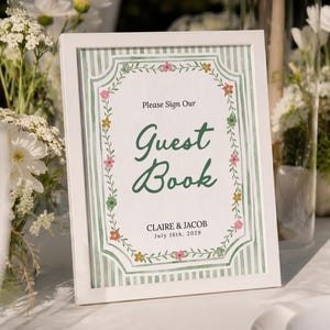 May include: A white framed sign featuring a green and white striped background with a floral border. The text reads: Please Sign Our Guest Book, CLAIRE & JACOB, July 16th, 2029. The design includes small colorful flowers in pink, yellow, and orange.