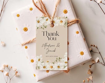 Op de afbeelding: Een cadeau verpakt in wit papier met madeliefpatronen, vastgebonden met touw, en een label met de tekst "Thank You Madison & Jacob SEP 24TH 2024" met bloemaccenten. Het label is beige met witte madeliefjes.