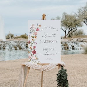 Editable “Love is in Bloom” bridal shower welcome sign template featuring a vertical border of soft watercolor wildflowers in pink, burgundy, lavender, and yellow, with modern script and serif fonts on a minimalist white background