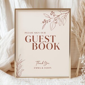 May include: A framed sign with the text "PLEASE SIGN OUR GUEST BOOK" in a serif font. The sign has a cream background with brown floral illustrations and the names "EMMA & JASON" at the bottom. The frame is gold.
