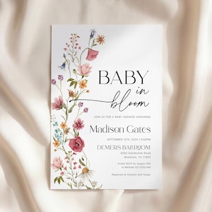 May include: A white baby shower invitation featuring floral designs. The text reads "BABY in bloom" with event details for Madison Gates, including the date, time, and location. The invitation is decorated with colorful flowers.