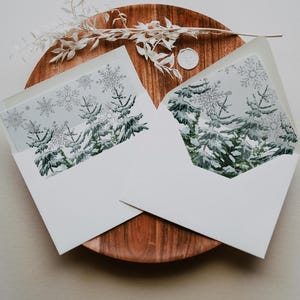 May include: Two white envelopes containing winter cards with snowy fir trees and snowflakes. The envelopes rest on a wooden tray, with a sprig of white flowers and a wax seal.