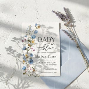 May include: A baby shower invitation with floral accents. The invitation features the words "BABY in bloom" in elegant script and includes details for Madison Gates' baby shower. A light blue envelope and lavender sprigs complete the scene.