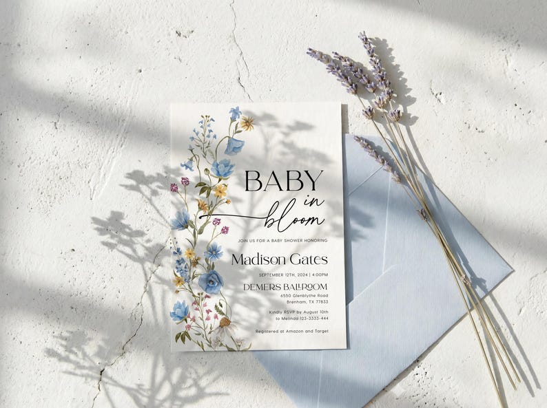 May include: A baby shower invitation with floral accents. The invitation features the words "BABY in bloom" in elegant script and includes details for Madison Gates' baby shower. A light blue envelope and lavender sprigs complete the scene.