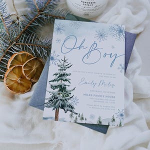 “Oh Boy” winter baby shower invitation with watercolor pine trees, delicate blue snowflakes, and elegant script text on a frosty blue background.
