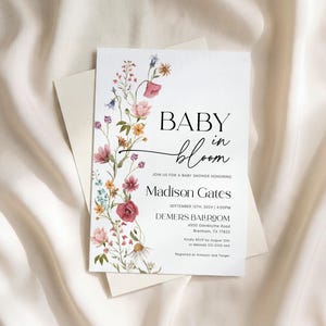 May include: A baby shower invitation featuring the words "BABY in bloom" in black script, adorned with watercolour flowers in pink, yellow, and blue. The invitation includes the name "Madison Gates" and event details.