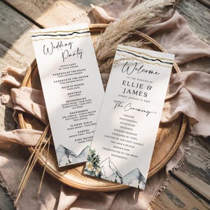 Op de afbeelding: Twee trouwceremonieprogramma's met een berg- en boomontwerp. De programma's hebben gouden en zwarte strepen aan de bovenkant en bevatten de details van de bruiloftsgasten en de volgorde van de ceremonie. De programma's liggen op een houten dienblad.