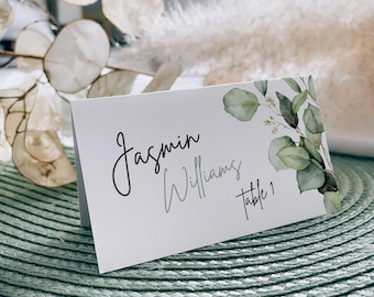 May include: White place card with green watercolor eucalyptus leaves and branches. The text reads "Jasmin Williams Table 1".