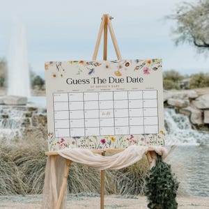 Könnte beinhalten: Eine weiße Karte mit einem floralen Rand und dem Text "Guess The Due Date of Sophie's Baby" oben. Die Karte hat ein Kalenderrraster mit den Zahlen 1-31 und den Wochentagen oben. Die Karte steht auf einem Holzständer.