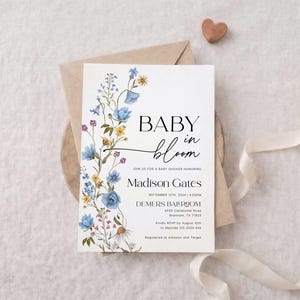 May include: A baby shower invitation with the words "BABY in bloom" in elegant script. The invitation features a floral design with blue, yellow, and purple flowers. The invitation is on a white background with a brown envelope and a ribbon.