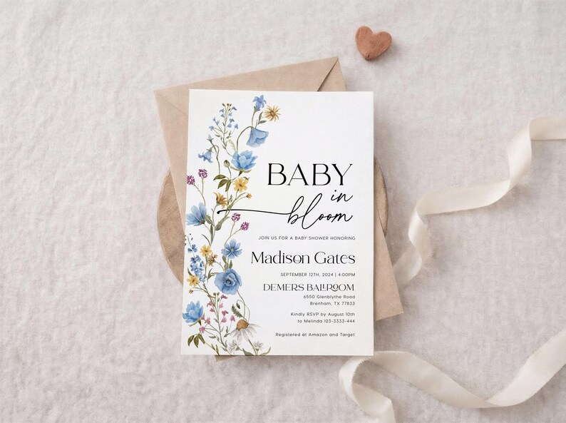 May include: A baby shower invitation with the words "BABY in bloom" in elegant script. The invitation features a floral design with blue, yellow, and purple flowers. The invitation is on a white background with a brown envelope and a ribbon.