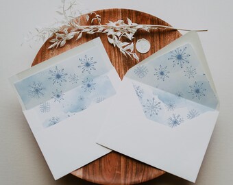 May include: Two white envelopes with blue snowflake designs inside, resting on a wooden plate. The envelopes are open, revealing the snowflake pattern. A sprig of white foliage and a wax seal are also on the plate, suggesting a winter or holiday theme.