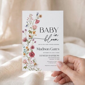 May include: A white baby shower invitation with the words "BABY in bloom" in black script. The invitation is decorated with a floral design in pink, purple, and yellow. The invitation includes the name Madison Gates and event details.