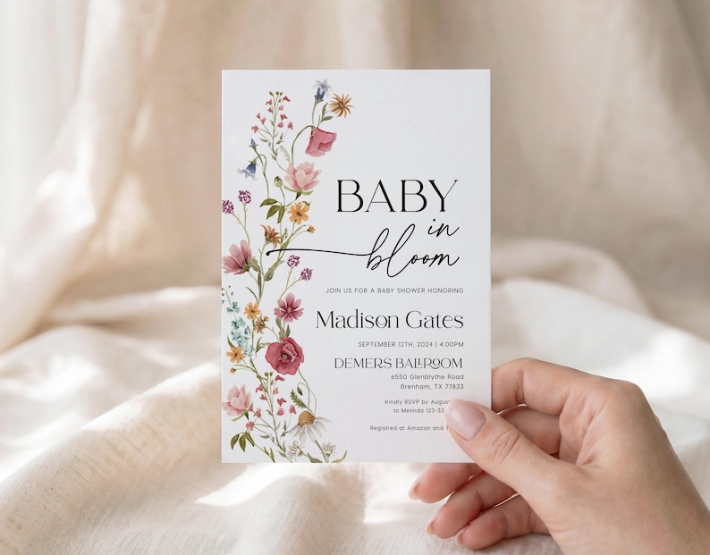 May include: A white baby shower invitation with the words "BABY in bloom" in black script. The invitation is decorated with a floral design in pink, purple, and yellow. The invitation includes the name Madison Gates and event details.