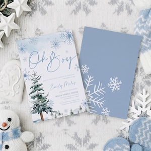May include: Two rectangular baby shower invitation cards featuring a winter theme. One card displays the text Oh Boy, a watercolor pine tree, and blue snowflakes. The second card is solid light blue with white snowflake illustrations on the corner.