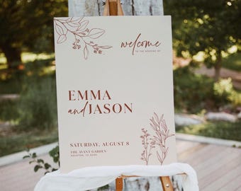 May include: A cream-colored wedding welcome sign on an easel. The sign features the names "EMMA and JASON" in a burgundy script, along with the date and location. Floral illustrations in burgundy accent the top and bottom corners.