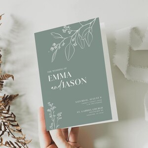 Può includere: Un biglietto d'invito di nozze piegato di colore verde tenue, con disegni floreali bianchi e il testo "The Wedding of Emma and Jason". La carta include anche la data, l'ora e il luogo della cerimonia. Un nastro bianco e una pianta secca sono sullo sfondo.