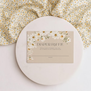 May include: A beige diaper raffle card with white daisy floral accents. The card reads "DIAPER RAFFLE" and includes space for a name. The card is on a white circular surface, with a floral fabric in the background.