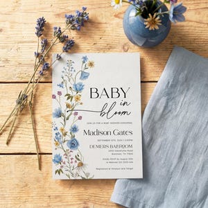 May include: A baby shower invitation with floral accents and the text "BABY in bloom." The invitation features blue and yellow flowers, with additional text including the name "Madison Gates" and event details. A blue napkin and vase with flowers are also present.