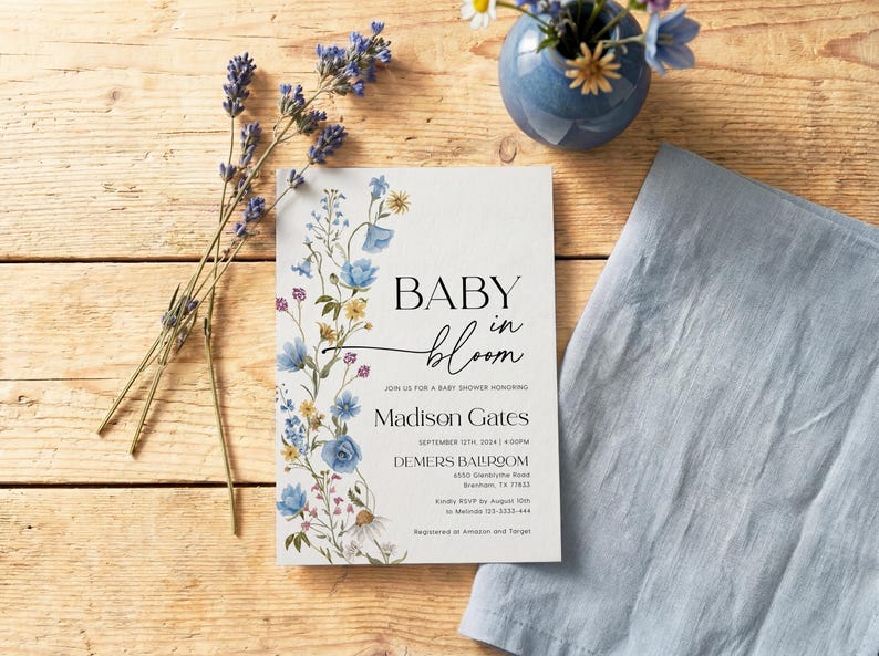 May include: A baby shower invitation with floral accents and the text "BABY in bloom." The invitation features blue and yellow flowers, with additional text including the name "Madison Gates" and event details. A blue napkin and vase with flowers are also present.