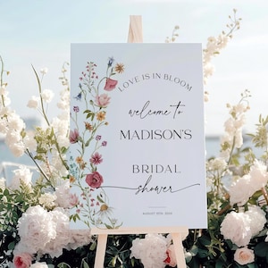 Editable “Love is in Bloom” bridal shower welcome sign template featuring a vertical border of soft watercolor wildflowers in pink, burgundy, lavender, and yellow, with modern script and serif fonts on a minimalist white background