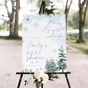 May include: A rectangular welcome sign for a baby shower featuring blue watercolor snowflakes, pine trees, and the text "Baby it's cold outside", "Welcome to Emily's baby shower", and "January 12, 2026". The sign is displayed on a black easel with white flowers.