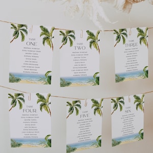 May include: Six white table cards with palm tree illustrations and guest names, suspended from a string. Each card features a beach scene at the bottom and the word "TABLE" followed by a number. The cards are held by silver clips.