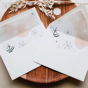 May include: Two white envelopes with snowflake and tree designs inside, resting on a wooden plate. The inner lining features a watercolor wash of pink and gray with silver snowflakes. A white wax seal and decorative sprig are also present.