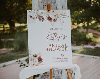 May include: A white sign on a wooden easel with the text "Welcome to Kelly's Bridal Shower" in brown script. The sign is decorated with floral arrangements in shades of white, brown, and beige. The date is September 12, 2023.