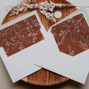 May include: Two white envelopes with a terracotta-colored interior featuring a delicate white botanical design. The envelopes are open, revealing the patterned interior. The envelopes rest on a wooden surface, with decorative elements.