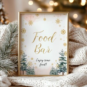 Op de afbeelding: Een ingelijst bord met gouden letters met de tekst "Food Bar" en "Enjoy some food!" op een witte achtergrond. Het bord is versierd met gouden sneeuwvlokken en roze accenten in aquarel. Kleine groenblijvende bomen staan onderaan.